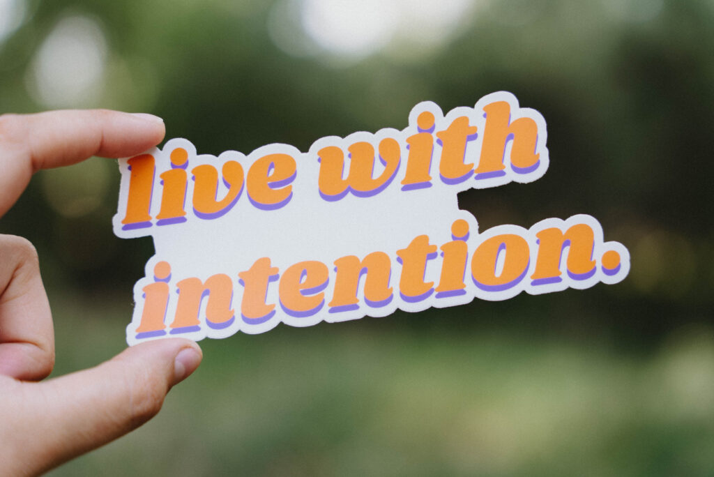 Spotlighting our Sticker Collection! Sticker that says "live with intention" held up in front of a green background