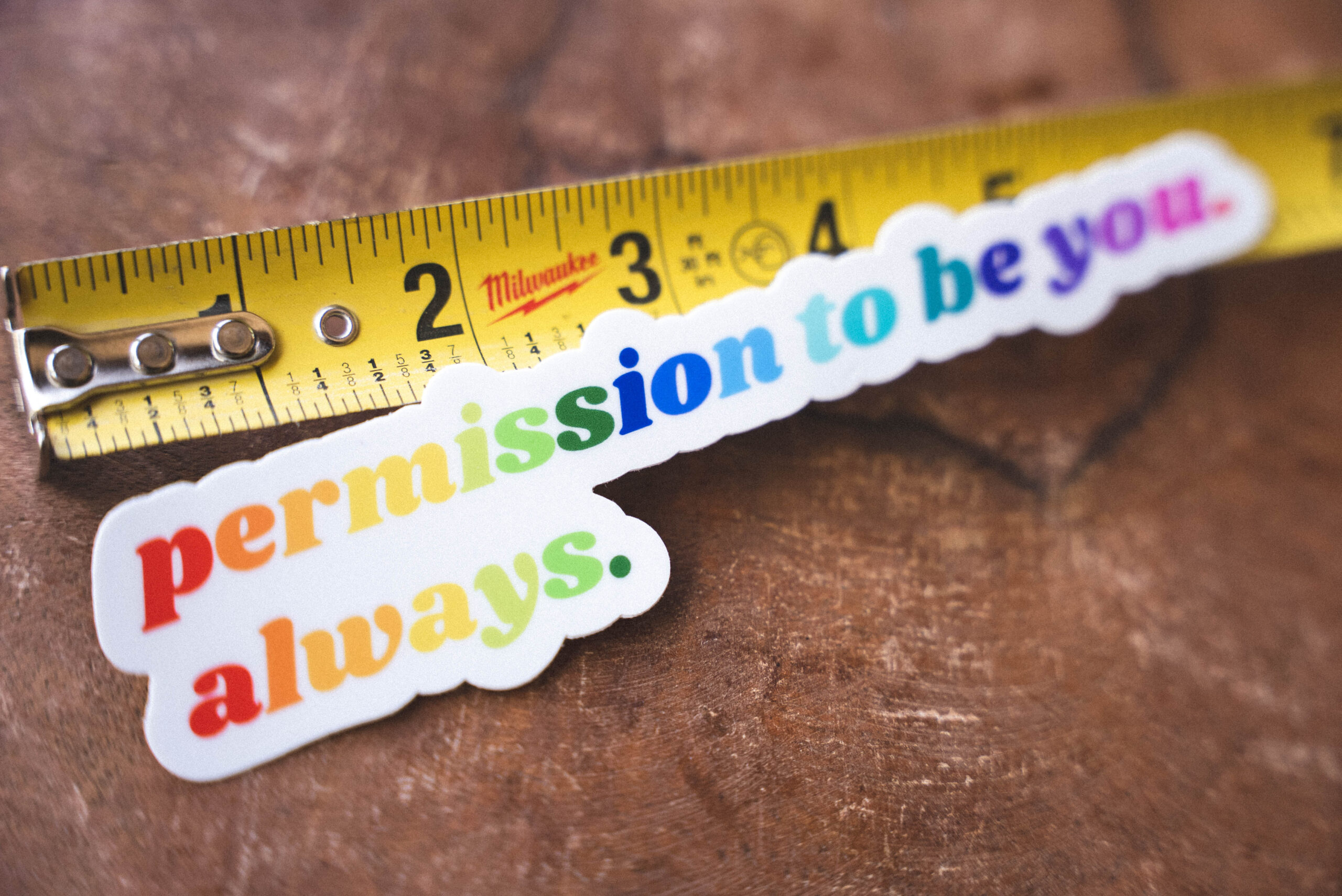 "Permission to be you. Always." Rainbow Sticker Sticker that says "permission to be you. always." and a tape measure on a table
