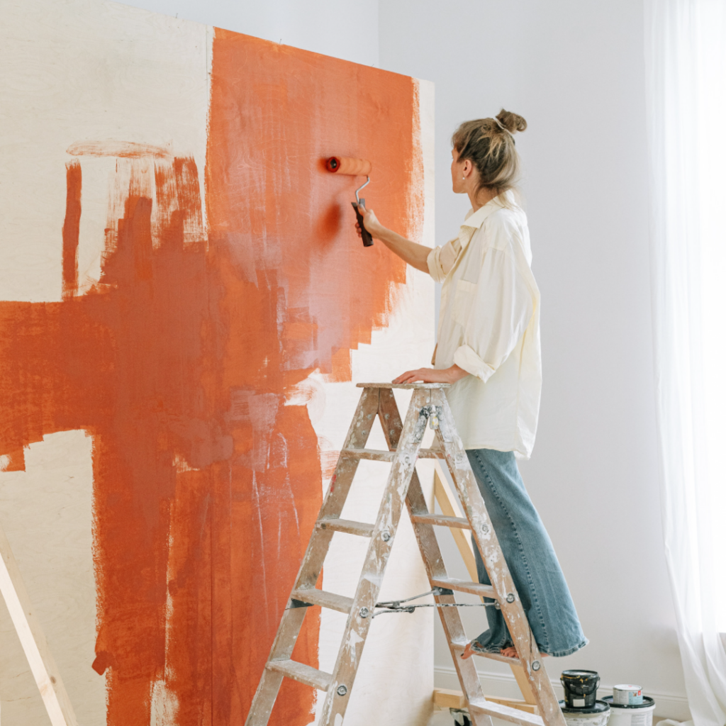 a woman on a ladder painting a white wall orange