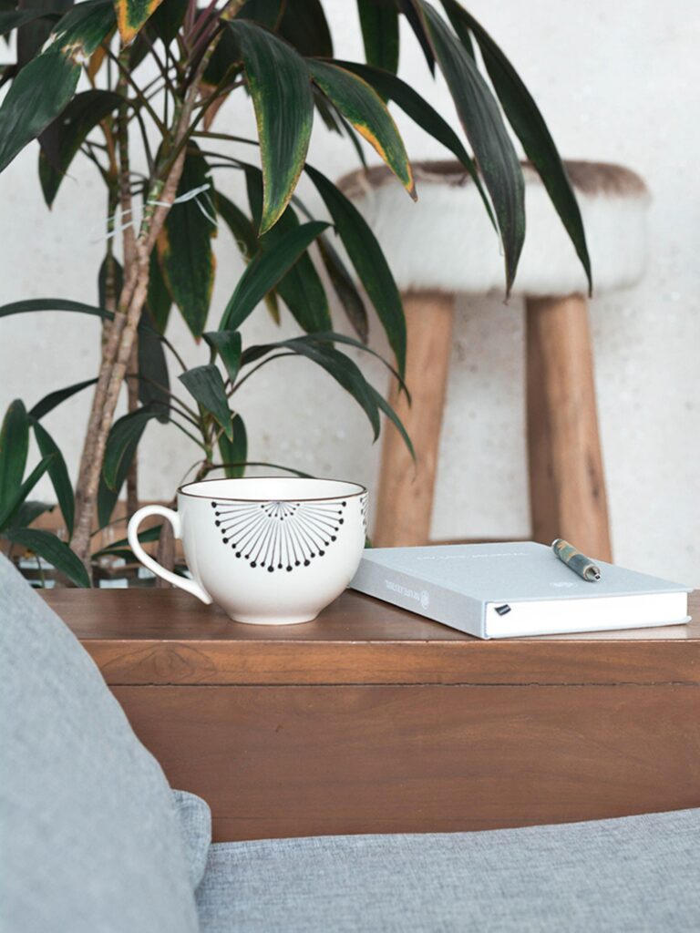 a mug and a journal and pen on a wooden table in front of a green plant